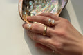 Hand wearing gold and pearl rings with an abalone shell in the background