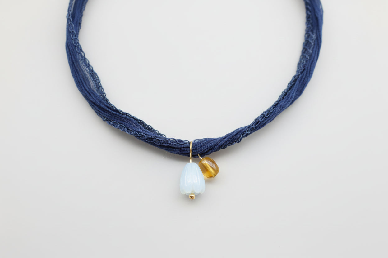 Denim blue silk ribbon necklace with a mop pikake pendant and amber nugget pendant on a light gray background by xomango